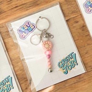 May include: A pink and silver keychain with a daisy charm and beaded accents. The keychain is attached to a card with the text "GREAT JOB!" and "BEST CLASS" in a colourful design.