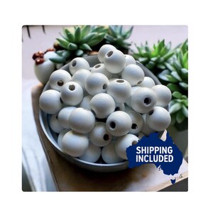May include: A collection of white wooden beads, each with a central hole, fills a light grey bowl. Green succulents are visible in the background. The image includes a blue graphic with the text "SHIPPING INCLUDED". The beads are ideal for crafting projects.