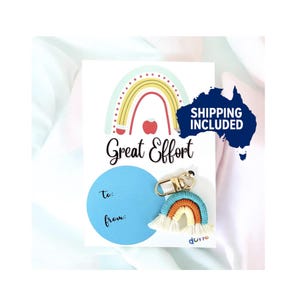 May include: A white card featuring a rainbow design and the words "Great Effort." A blue circle with "to:" and "from:" is below the text. A small rainbow keychain with gold hardware is attached to the card.