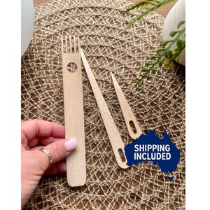 May include: Three wooden weaving tools: a weaving fork and two weaving needles. The tools are light-coloured wood and are arranged on a textured, brown surface. The image includes a hand with a gold ring.