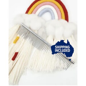 May include: A colourful rainbow wall hanging with a metal pet grooming comb. The rainbow has red, yellow, pink, and blue arches with fluffy white cloud accents. The comb is silver and has closely spaced metal teeth.