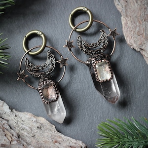 May include: Two crystal quartz earrings with bronze-colored accents. Each earring features a clear quartz crystal pendant, a crescent moon, and star details. The earrings are suspended from a bronze-colored hoop.