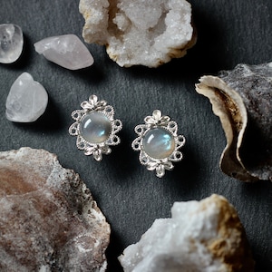 May include: A pair of silver earrings with a delicate floral design and iridescent moonstone cabochons. The earrings are displayed on a dark background with various crystals and shells.