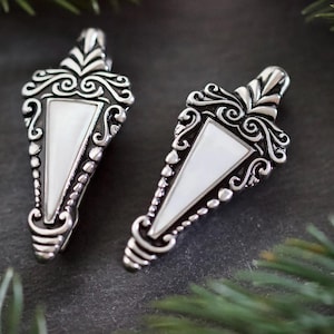 May include: Two silver-toned earrings with a triangular white inlay and ornate scrollwork. The earrings have a teardrop shape with a decorative top and a beaded border. The background is dark with green foliage.
