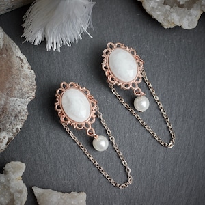 May include: A pair of elegant earrings featuring oval white cabochons set in ornate rose gold frames. Each earring has a delicate chain and a dangling pearl. The jewelry is displayed on a dark gray surface.