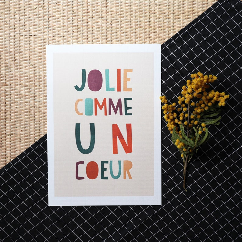 May include: A beige card with the text 'JOLIE COMME UN COEUR' in colorful, stylized letters. The letters are in various colors including red, orange, yellow, green, blue, and purple.