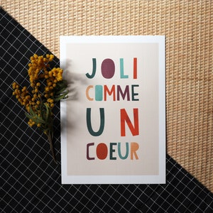May include: A white card with the text 'JOLI COMME UN COEUR' in colorful letters. The letters are in different colors, including red, orange, yellow, green, blue, and purple. The card is on a woven background.