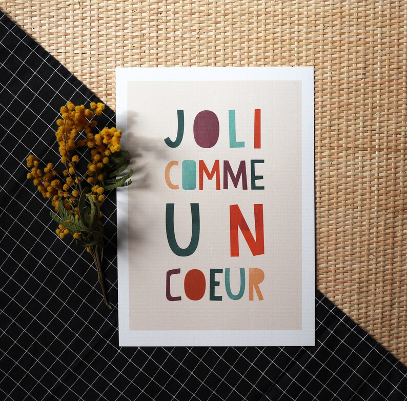 May include: A white card with the text 'JOLI COMME UN COEUR' in colorful letters. The letters are in different colors, including red, orange, yellow, green, blue, and purple. The card is on a woven background.