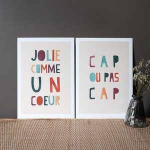 May include: Two framed prints with colorful text. The print on the left has the text 'Jolie comme un coeur' and the print on the right has the text 'Cap ou pas cap'.