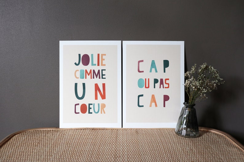 May include: Two framed prints with colorful text. The print on the left has the text 'Jolie comme un coeur' and the print on the right has the text 'Cap ou pas cap'.