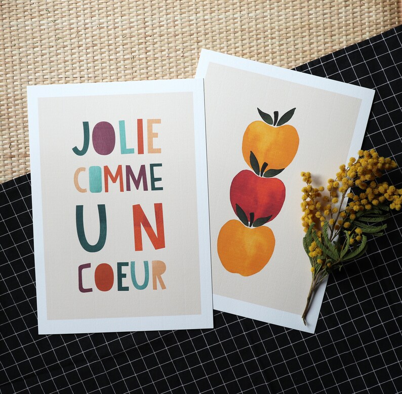 May include: Two cards with a white background and colorful text. The card on the left has the text 'JOLIE COMME UN COEUR'. The card on the right has a stack of three apples. The top apple is yellow, the middle apple is red, and the bottom apple is yellow. The apples are stacked on top of each other. There is a small branch of yellow flowers in the corner of the card on the right.