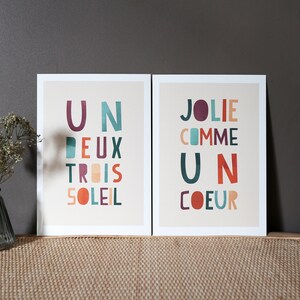 May include: Two framed prints with colorful text on a beige background. The print on the left says 'Un Deux Trois Soleil' and the print on the right says 'Jolie Comme Un Coeur.'