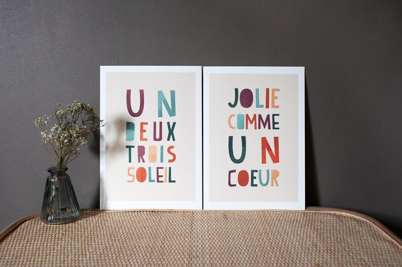May include: Two framed prints with colorful text on a beige background. The print on the left says 'Un Deux Trois Soleil' and the print on the right says 'Jolie Comme Un Coeur.'