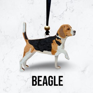 May include: Wooden ornament of a beagle dog with black, brown, and white markings. The ornament is hanging from a black ribbon with a gold bead.