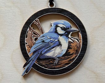 Op de afbeelding: Een blauwe gaaien ornament met een zwarte rand en een bruin nestontwerp. Het ornament is gemaakt van hout en heeft een touwtje om op te hangen.