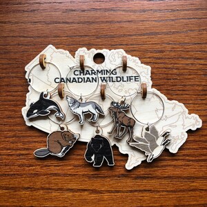 May include: Set of six wine glass charms featuring Canadian wildlife. The charms are made of wood and feature a moose, wolf, beaver, bear, orca, and goose. The charms are attached to a wooden map of Canada with the text "Charming Canadian Wildlife".