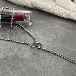 May include: A silver-toned necklace with a linear pendant and a chain design. The necklace is displayed on a gray surface next to a tube of lipstick with the words "NON-STOP INTENSE COLOR!"