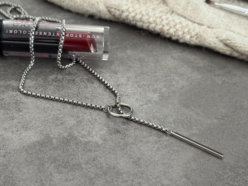 May include: A silver-toned necklace with a linear pendant and a chain design. The necklace is displayed on a gray surface next to a tube of lipstick with the words "NON-STOP INTENSE COLOR!"