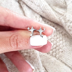 Op de afbeelding: Zilveren stervormige oorbellen met een cluster van vijf sterren. De oorbellen worden getoond op een hand met witte nagellak.