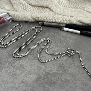 May include: A silver-toned necklace with a bar pendant and a chain-link design. The necklace is displayed on a gray surface, alongside makeup brushes and a tube of lipstick. A cream-colored knitted item is in the background.