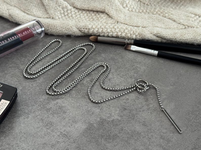 May include: A silver-toned necklace with a bar pendant and a chain-link design. The necklace is displayed on a gray surface, alongside makeup brushes and a tube of lipstick. A cream-colored knitted item is in the background.