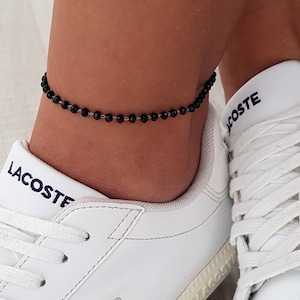 Può includere: Un braccialetto da caviglia con perline nere sul piede di una persona. Il braccialetto è fatto di piccole perline rotonde che sono collegate da una catena sottile. La persona indossa scarpe da ginnastica bianche con la scritta "LACOSTE" sul lato.