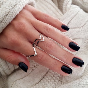 Peut inclure: Une bague en argent avec un motif en chevron. La bague est portée sur le majeur d'une main avec un vernis à ongles noir.