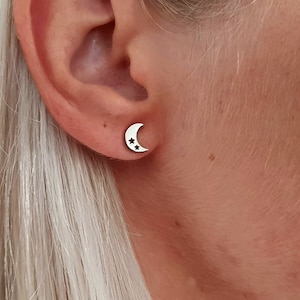 Può includere: Un paio di orecchini a forma di mezzaluna in argento con due piccole stelle ritagliate dalla luna. Gli orecchini sono mostrati nell'orecchio di una persona.