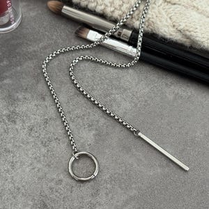 May include: A silver-colored necklace with a chain and a bar pendant. The necklace features a circular ring at the end of the chain. The jewelry is displayed on a gray surface, with makeup brushes and a lipstick tube in the background.