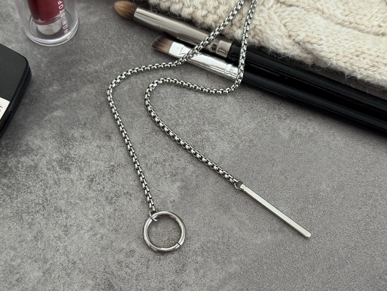 May include: A silver-colored necklace with a chain and a bar pendant. The necklace features a circular ring at the end of the chain. The jewelry is displayed on a gray surface, with makeup brushes and a lipstick tube in the background.