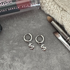 Può includere: Un paio di orecchini a cerchio argentati con ciondoli a forma di simbolo del dollaro. Pennelli per il trucco, rossetto e un articolo lavorato a maglia sono visibili su una superficie grigia. Il rossetto recita "NON-STOP INTENSE COLOR!"