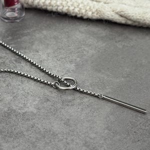 May include: A silver-toned necklace with a delicate chain and a linear bar pendant. The necklace features a small ring detail connecting the chain to the bar. The jewelry is displayed on a gray surface.