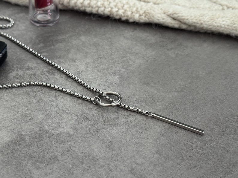 May include: A silver-toned necklace with a delicate chain and a linear bar pendant. The necklace features a small ring detail connecting the chain to the bar. The jewelry is displayed on a gray surface.