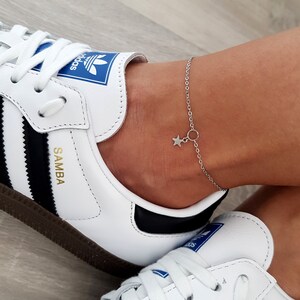 Peut inclure: Un bracelet de cheville argenté avec un charme en forme d'étoile et un petit charme rond. Le bracelet est sur la cheville d'une personne, et la personne porte des baskets blanches avec des rayures noires et le mot "SAMBA" écrit sur le côté.