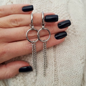 Pode incluir: Um par de brincos de argola prateados com correntes penduradas. Os brincos s&atilde;o segurados em uma m&atilde;o com esmalte de unha preto.