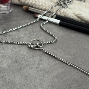 May include: A silver-colored necklace with a circular pendant and a vertical bar pendant. The necklace chain has a box link design. Makeup brushes and a cosmetic container are in the background.