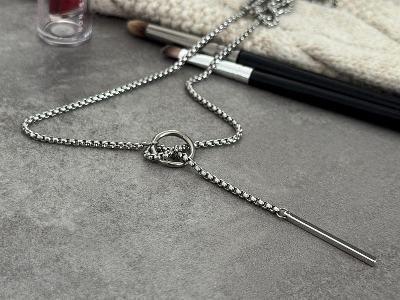 May include: A silver-colored necklace with a circular pendant and a vertical bar pendant. The necklace chain has a box link design. Makeup brushes and a cosmetic container are in the background.