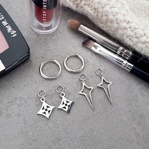 Könnte beinhalten: Ein Set silberner Ohrringe und Charms. Die Ohrringe sind hufeisenförmig und die Charms sind ein Stern und ein geometrisches Design.