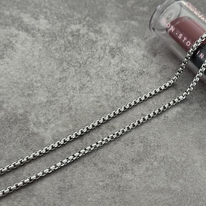 May include: A silver-colored box chain necklace. The necklace is made of small, square links. A tube of lipstick is in the background. The lipstick tube is black with white text and a red lipstick bullet.