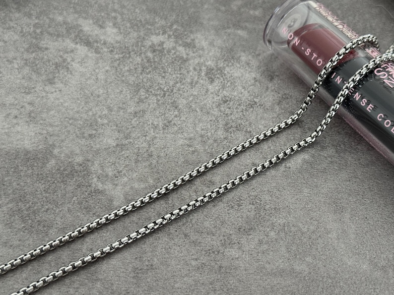 May include: A silver-colored box chain necklace. The necklace is made of small, square links. A tube of lipstick is in the background. The lipstick tube is black with white text and a red lipstick bullet.