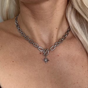 Könnte beinhalten: Eine silberne Kettenkette mit einem sternförmigen Anhänger. Die Kette besteht aus dickem, verlinktem Metall und hat einen Toggle-Verschluss.