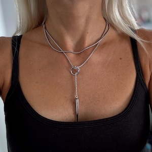 Può includere: Una collana argentata con un design a strati. Lo strato superiore è una catena delicata, mentre lo strato inferiore presenta un anello e un lungo pendente a punta. La collana è indossata su un top nero.