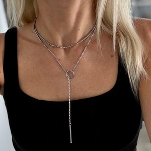 May include: A silver-colored necklace with two strands. The top strand is a delicate chain, while the lower strand features a circular pendant with a vertical bar. The necklace is worn against a black top.