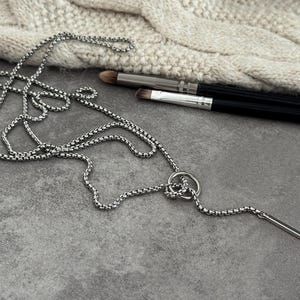 May include: A silver-toned necklace with a bar pendant and a circular accent. The necklace is displayed on a gray surface with makeup brushes and a tube of lipstick in the background. The chain has a box link design.