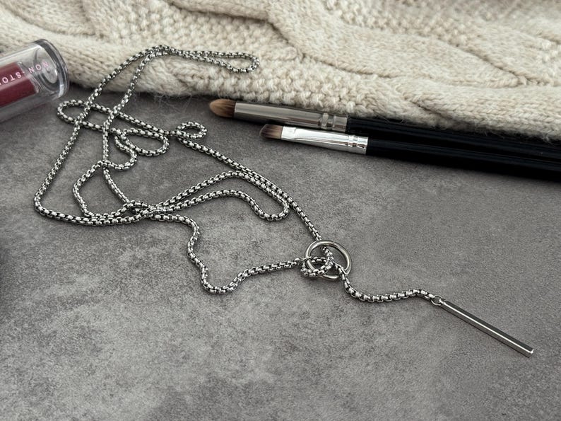 May include: A silver-toned necklace with a bar pendant and a circular accent. The necklace is displayed on a gray surface with makeup brushes and a tube of lipstick in the background. The chain has a box link design.