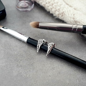 May include: A pair of silver angel wing earrings with a delicate, intricate design. The earrings are shown on a black makeup brush.