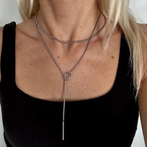 May include: A silver-colored necklace with a layered design. The necklace features a delicate chain that forms a loop, with a vertical bar pendant. The necklace is worn against a black top.