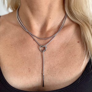 May include: A silver-toned necklace with a layered design. The necklace features two strands of small, round beads, with a circular pendant and a vertical bar pendant. The necklace is worn against a black top.