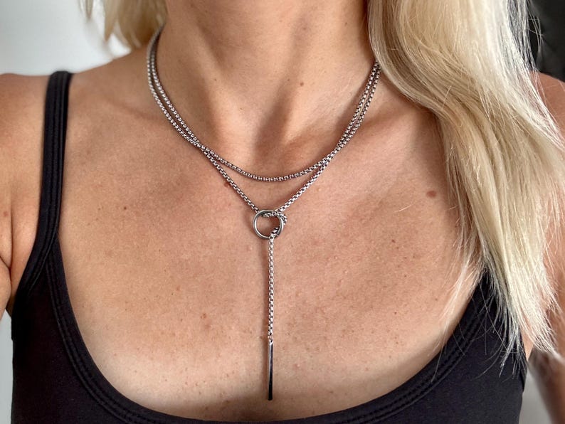 May include: A silver-toned necklace with a layered design. The necklace features two strands of small, round beads, with a circular pendant and a vertical bar pendant. The necklace is worn against a black top.