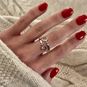 Könnte beinhalten: Ein silberner Ring mit zwei verbundenen Bändern, die jeweils mit einem stilisierten Frauensymbol verziert sind. Der Ring wird an einem Finger mit rotem Nagellack getragen. Der Hintergrund ist ein cremefarbener Zopfstrickpullover.